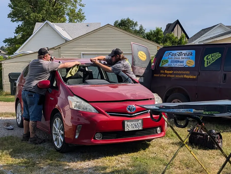 Two Fas-Break technicians working on a red Toyota Prius windshield in a driveway, next to the maroon Fas-Break van